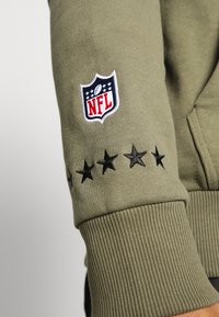 La manche du sweatshirt vert olive présente un patch du logo de la NFL et des étoiles noires brodées. La texture côtelée du poignet est visible, créant un look ajust é.