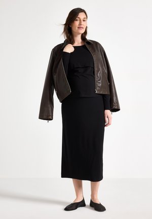 Femme portant une robe noire et une veste en cuir marron drapée sur les épaules, debout devant un fond blanc uni.