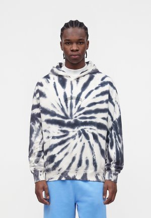 Młody mężczyzna z plecionymi włosami, ubrany w czarno-białą bluzę tie-dye z napisem "Jordan" oraz jasnoniebieskie spodnie, stoi zwrócony twarzą do przodu.