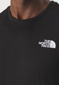 Pohled zblízka na černé tričko s kulatým výstřihem, na kterém je bílé logo "The North Face" na levé straně hrudi, které nosí osoba.