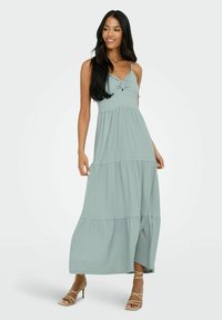 Robe maxi de couleur vert clair, avec de fines bretelles, un détail torsadé au niveau du bustier et une jupe à volants. Texture lisse, silhouette fluide, longueur cheville.