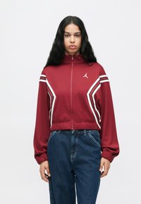 Kobieta o długich ciemnych włosach nosi bordową kurtkę z zamkiem błyskawicznym z białymi i czarnymi paskami oraz luźne niebieskie dżinsy na jednolitym tle.