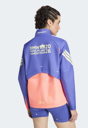 Person med krøllet hår bærer en blå og koraljakke med BMW Berlin Marathon 2026-logo og matchende blå shorts, vist bagfra.