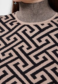 Maglione lavorato a maglia con colletto alto a coste, caratterizzato da un motivo geometrico nero su una base beige morbida, con attenzione alla texture e ai dettagli del design.