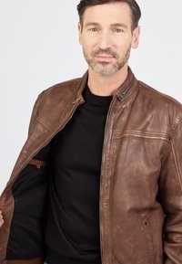 Bruine leren jas met een ritssluiting aan de voorkant, staande kraag en horizontale stiksels over de borst. Draagt over een zwarte trui.