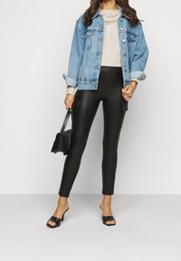 Spijkerjack, lichtblauw, losse pasvorm, met knoopsluiting aan de voorkant. Beige trui eronder, zwarte broek van nep leer met zijzakken. Zwarte tas, openhakken.