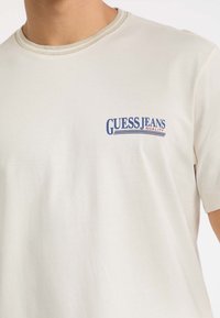 Zbliżenie na osobę w off-white koszulce z krótkim rękawem z niebieskim i czerwonym logo "GUESS JEANS QUALITY" na lewej piersi.