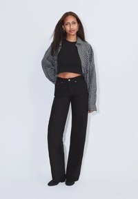 Frau mit langen Haaren, die ein schwarzes Crop-Top, ein übergroßes kariertes Hemd und hoch taillierte schwarze Hosen trägt, steht vor einem schlichten hellen Hintergrund.