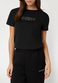 Juoda T-maikė, pagaminta iš minkštos medžiagos, su apvali apykakle ir blizgiuoju "GUESS" logotipu priekyje. Dėvima su derančiomis juodomis kelnėmis.