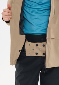 Beige wasserdichte Jacke mit durchgehendem Reißverschluss. Die Innenseite zeigt ein blaues Strukturhemd und einen schwarzen Bund mit Druckknopfverschlüssen und elastischem Material.