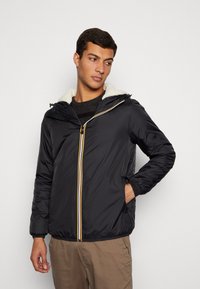 Giacca imbottita nera con cappuccio, dotata di una fodera in pile crema e una chiusura frontale con cerniera arricchita da accenti gialli e bianchi. Vestibilità casual con tasche laterali.