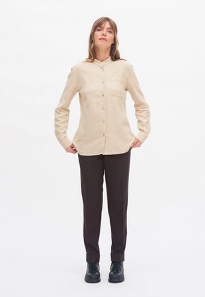 Woman standing wearing beige button-up shirt, dark brown pants, and black lace-up shoes against a plain white background.