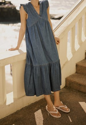 Vrouw in mouwloze blauwe denim midi-jurk en witte teenslippers leunend op een crème kleurige reling bij een door de zon verlichte buitentrap.