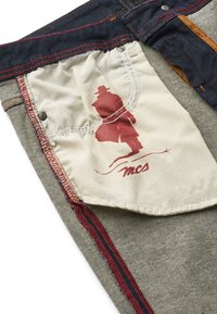 Zbliżenie podszewki kieszeni spodni jeansowych z czerwonym konturem osoby w kapeluszu i płaszczu nad tekstowym logiem „mcs“.