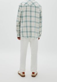 Chemise à carreaux beige, bleue et verte, à manches longues avec poignets à boutons, associée à un pantalon blanc et des baskets bicolores.