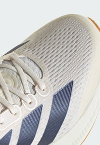 Ľahká bežecká obuv s textúrovaným bielym sieťovinovým zvrškom, námořníckou modrou pruhovanou akcentáciou a gumovou podrážkou. Šnúrky majú dizajn s slučkami.