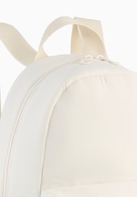 Un sac à dos beige clair avec une forme arrondie, un tissu lisse, des fermetures éclair et des bretelles robustes. Il présente une texture subtile et un design minimaliste.