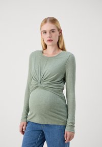 Hosszú ujjú, bordás kismama felső lágy zöld színben, csavart elülső dizájnnal és kerek nyakkivágással. Kék farmernadrággal párosítva.