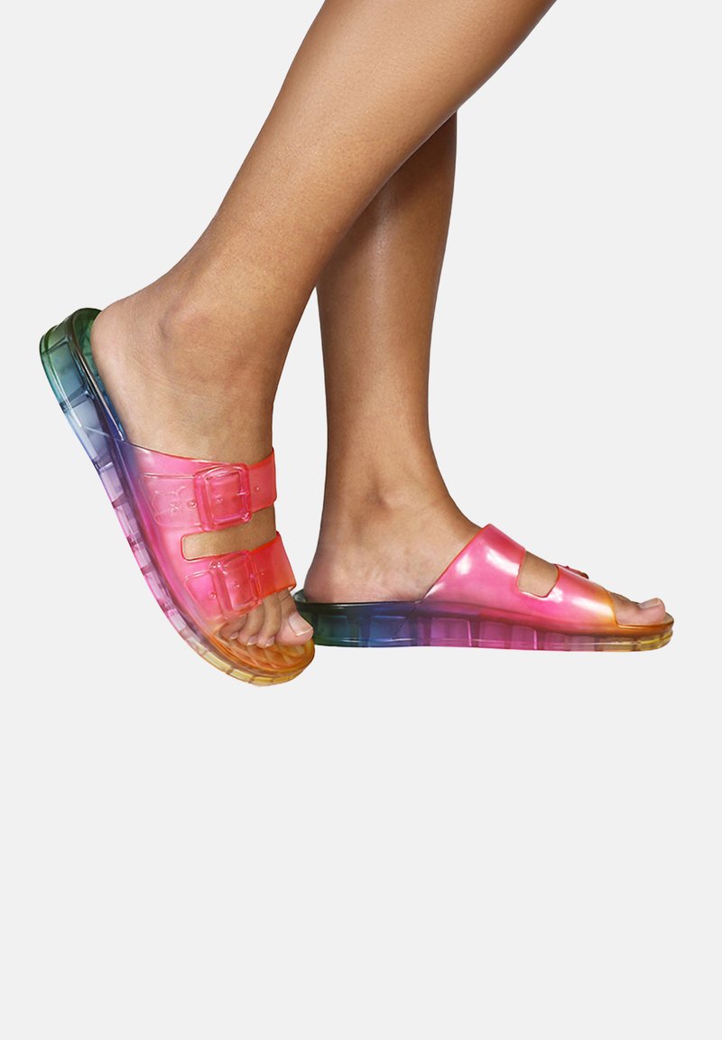 Persona che indossa sandali trasparenti color arcobaleno con doppie fibbie, mostrando i piedi e le gambe inferiori su uno sfondo bianco.