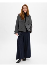 Grijze wollen peacoat met dubbelrijige knopen, gecombineerd met marineblauwe wijde broek. Model houdt een getextureerde clutch vast en draagt zwarte hakken.