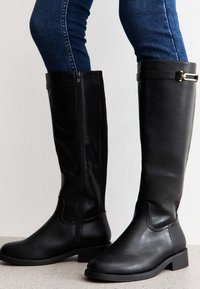 Bottes noires en cuir montant jusqu'au genou avec une fermeture éclair sur le côté et une petite sangle décorative avec un accent doré. Talon bloc robuste. Texture lisse.