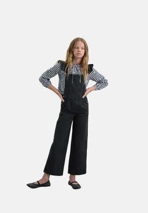 Pantalones de tirantes negros de pierna ancha llevados sobre una blusa a cuadros con hombros con volantes y un escote con cordón. Zapatos planos negros con remaches.