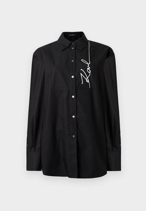Chemise noire à manches longues avec col et broderie cursive blanche "Karl" sur la partie droite de la poitrine.