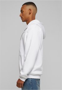 White zip-up hoodie made from soft fabric, featuring a large kangaroo pocket, adjustable hood, and ribbed cuffs, paired with blue jeans.