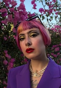 Paarse blazer, korte roze en blonde haren, gedurfde zwarte eyeliner met goud, rode lippen, parelketting en bloemenachtergrond.