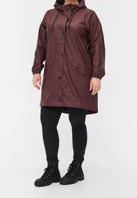 Bordeaux nepromokavý plášť s kapucí, přední zapínání na patentky, elastické manžety a dvě přední kapsy, nošený přes černé kalhoty a boty.
