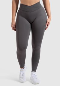 Leggings grises de talle alto, confeccionados en una tela texturizada y elástica. Presentan una cinturilla cruzada y un ajuste elegante. Llevados con zapatillas blancas.
