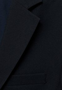 Närbild av en mörk marinblå kavajlapp och bröstficka som är gjord av texturerad tyg.