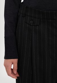 Mustade triibulised püksid voldikutega, esiküljel nupuga kinnitamine ja struktureeritud vöökoht. Kanga tekstuur on viimistletud ja kohandatud sobivuse jaoks.