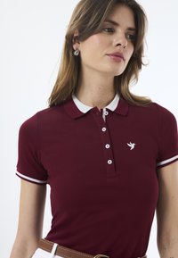 Burgundia polo särk valgete äärtega, millel on krae ja viis nuppe. Rinnas on väike tikitud linnulogo.