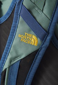 Chaleco de North Face en teal y malla negra, con correas azules y un logotipo amarillo de diseño curvado. Tela texturizada y detalles de costura visibles.