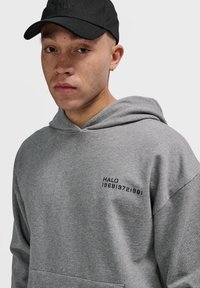 Młody mężczyzna z jasnymi piegami, noszący czarną czapkę i szary hoodie z nadrukiem "HALO 196819721981" na klatce piersiowej.