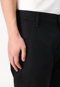 Juodi kelnių kelnės pagamintos iš medvilnės mišinio, su siauru siluetu, šoninėmis kišenėmis ir glotnia tekstūra, turinčiomis tvirtą juosmenį.