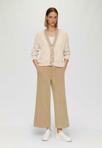 Beige Strickjacke mit einem kontrastierenden braunen Kragen, Knopfverschluss, weiten weißen Hosen und weißen Sneakers, vor einem neutralen Hintergrund.