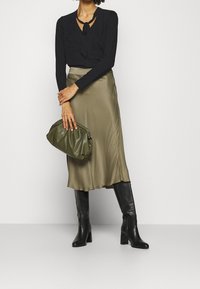 Svart långärmad topp med slingdetalj, olivgrön satin midi-kjol, svarta knähöga klackstövlar och en grön klutch. Släta texturer och åtsittande former.