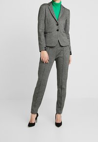 Costume texturé gris avec une veste ajustée et un pantalon sur mesure, associé à un col roulé vert et des chaussures à talons noirs.