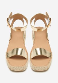 Sandalias espadrilles metálicas doradas con una correa para el tobillo, suela de yute trenzado y una planta de cuero suave. Presenta un diseño plano y abierto en la parte delantera.