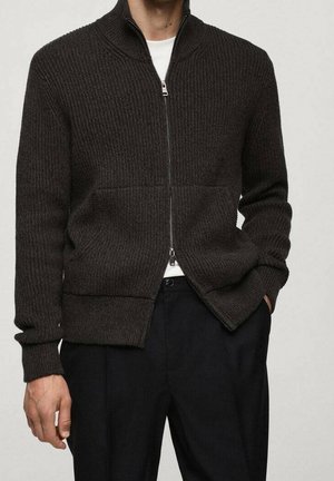 Mężczyzna ubrany w ciemnobrązowy sweter na zamek z dzianiny, na biały t-shirt, w czarnych spodniach, stoi z jedną ręką w kieszeni.