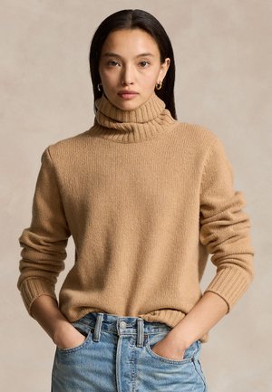 Woman wearing a beige turtleneck sweater and light blue jeans with hands in pockets against a plain background.