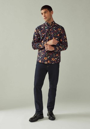 Knopjesoverhemd van marineblauwe stof met een kleurrijk patroon van voedsel en ruimte; gecombineerd met donkere jeans en zwarte schoenen.