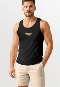 Black tank top with a rounded neckline and light gold logo in the center, paired with beige shorts, showcasing a fitted design.