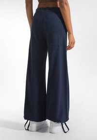 Marineblauwe wijde broek met een zwarte merkband, twee achterzakken en een rechte textuur. Gestyled met witte sneakers.