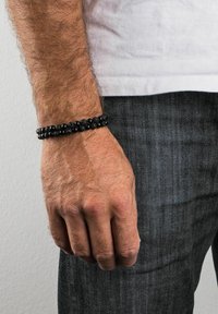 Melnā pērlīšu aproce, kas izgatavota no apaļām akmens pērlītēm, ar gludu, spīdīgu virsmu. Valkāta uz rokas ar gaišiem matiem, uz pelēka fona.