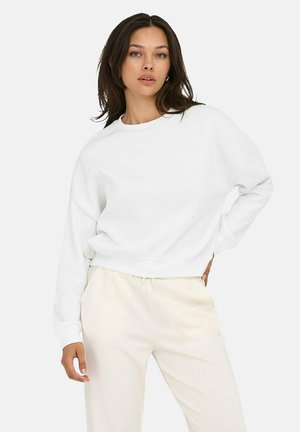 Felpa bianca in cotone con scollo rotondo, spalle drop e polsini a costine, abbinata a pantaloni beige chiaro con cordino.