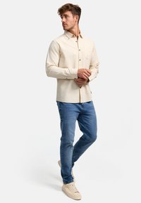 Chemise crème à manches longues avec un tissu texturé et des détails de boutons, assortie à un jean bleu clair et des baskets beiges.