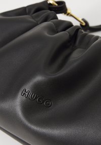 Borsa in pelle nera con parte superiore arricciata, caratterizzata dal logo "HUGO" in rilievo e dettagli in oro. Texture liscia e forma curva.